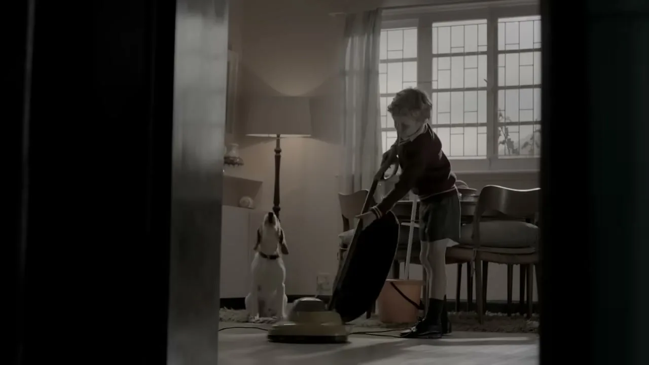 A young boy in a red sweater and shorts vacuums a living room while a beagle dog sits patiently nearby, looking up at him.