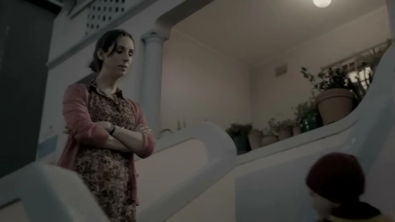 A pregnant woman with her arms crossed looks down at a small child in a red hat and yellow jacket on a staircase in a house.