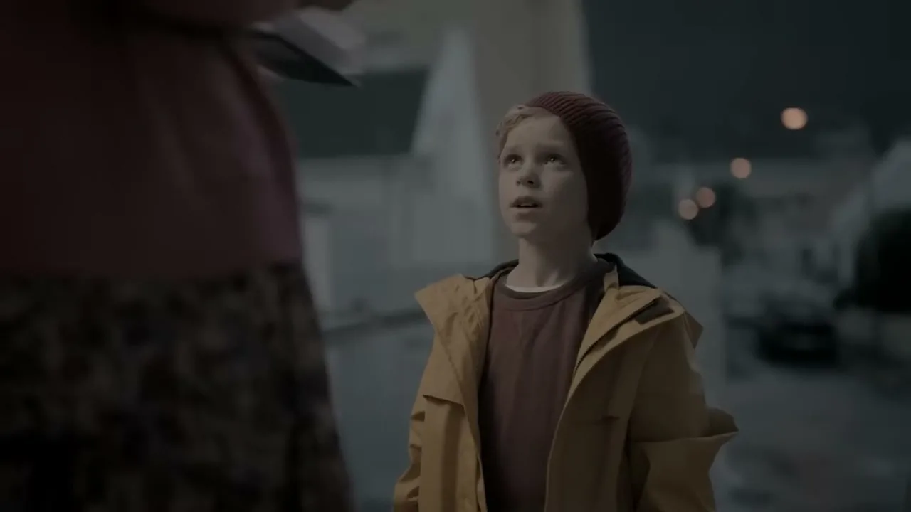 A young boy in a yellow jacket and red beanie looks up at an adult, standing on a street at night.