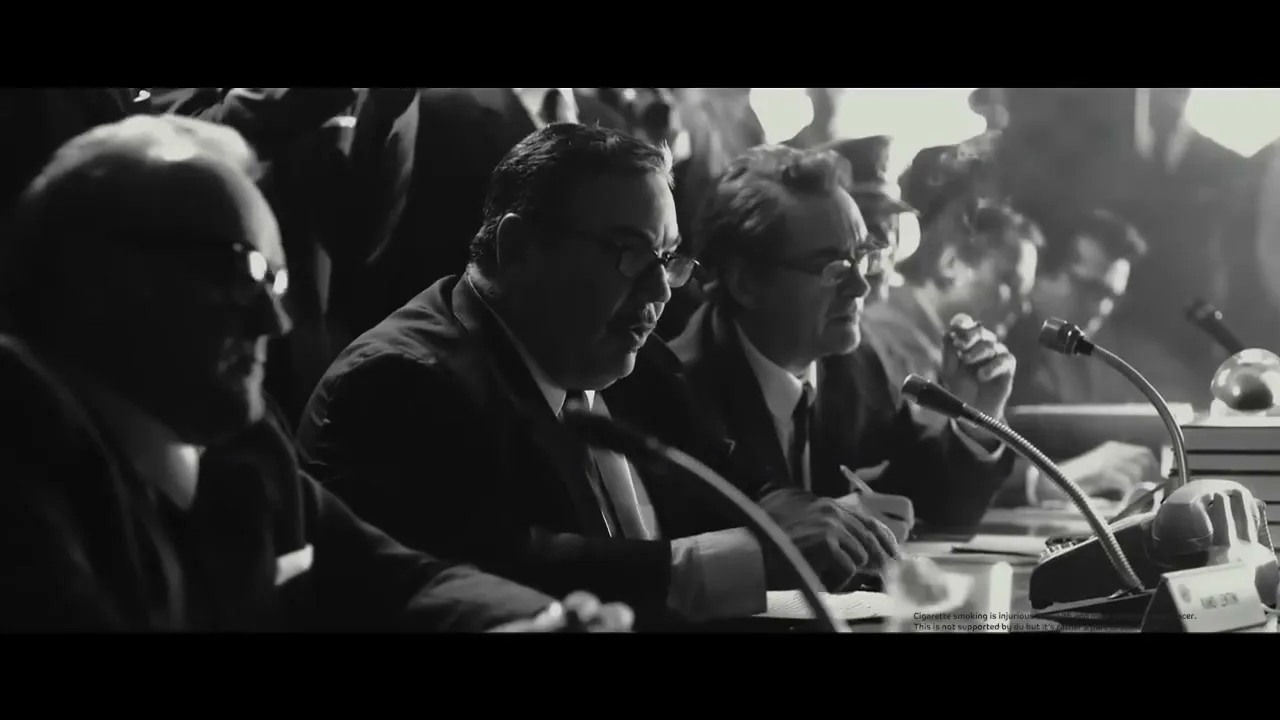 A black and white shot shows several men in suits sitting at a long table with microphones, appearing to be at a press conference or meeting.