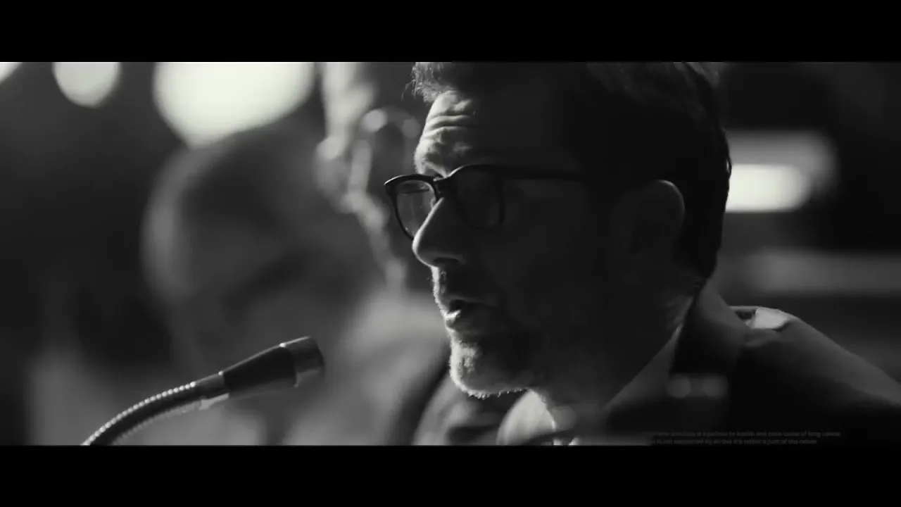 A man with glasses and a beard speaks into a microphone at a formal event, with another person blurred in the background.