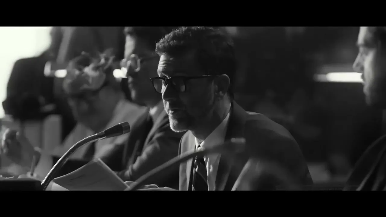 A man in his 40s, wearing glasses and a suit, speaks into a microphone at a formal hearing, with other people blurred in the background.