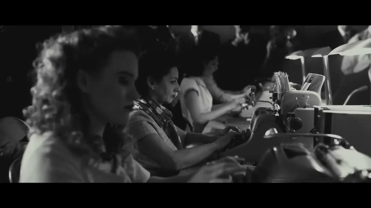 A group of women in a 1940s office are typing on typewriters, focused on their work in a dimly lit room.