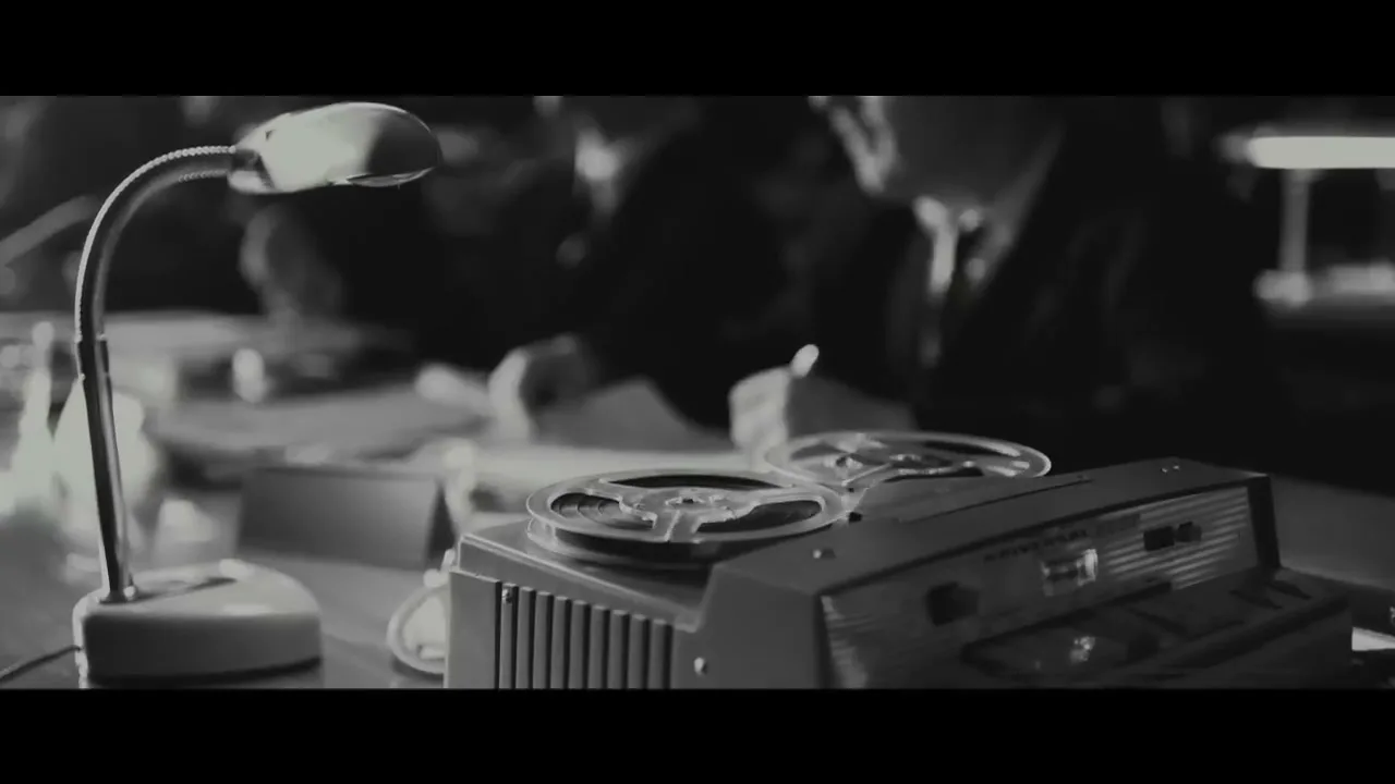 A black and white shot shows a vintage reel-to-reel tape recorder and a desk lamp in the foreground, with a man writing at a desk in the background.