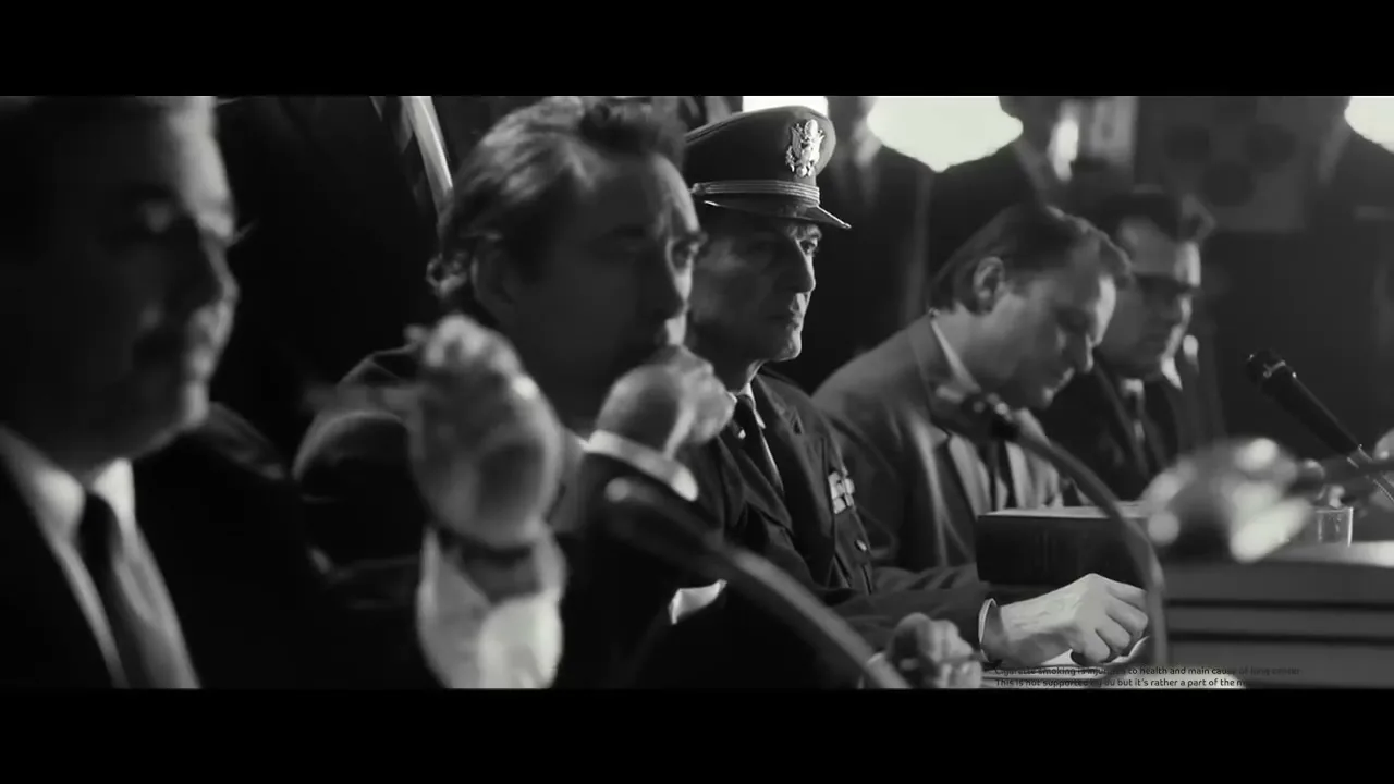 A black and white shot shows several men in suits and one in a military uniform sitting at a long table with microphones, appearing to be at a formal hearing or conference.