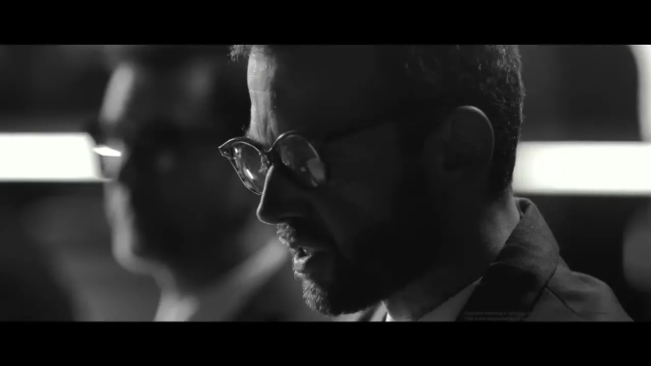 A man with glasses and a beard, wearing a suit, adjusts his glasses while speaking in a dimly lit interior setting.