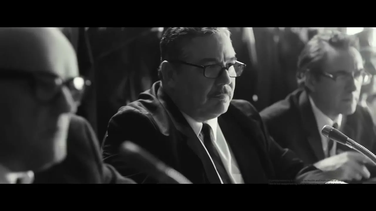 A man with glasses and a mustache sits at a table with other men, looking serious in a black and white, low-key setting.