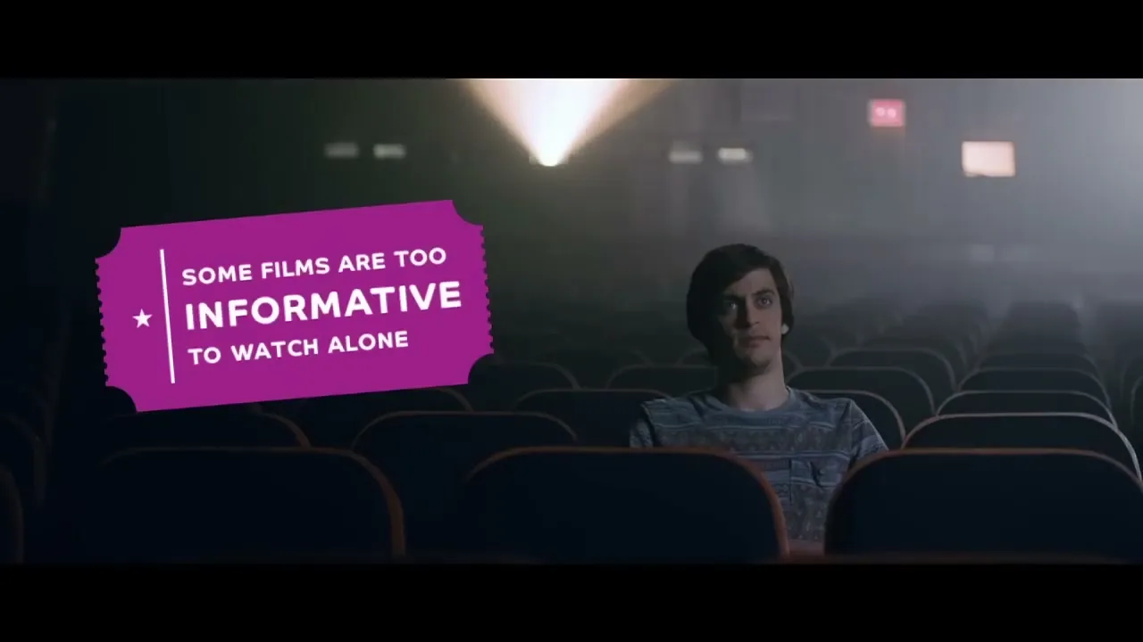 A young man with dark hair sits alone in a dark, empty movie theater, looking forward as a projector beam shines from behind him.