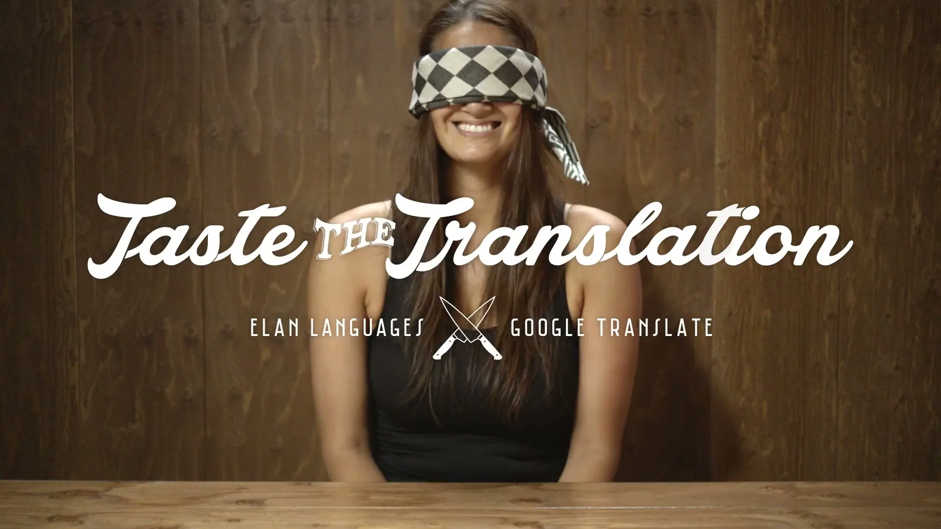 A blindfolded woman with a checkered blindfold sits at a wooden table, smiling, with text overlayed on the screen.