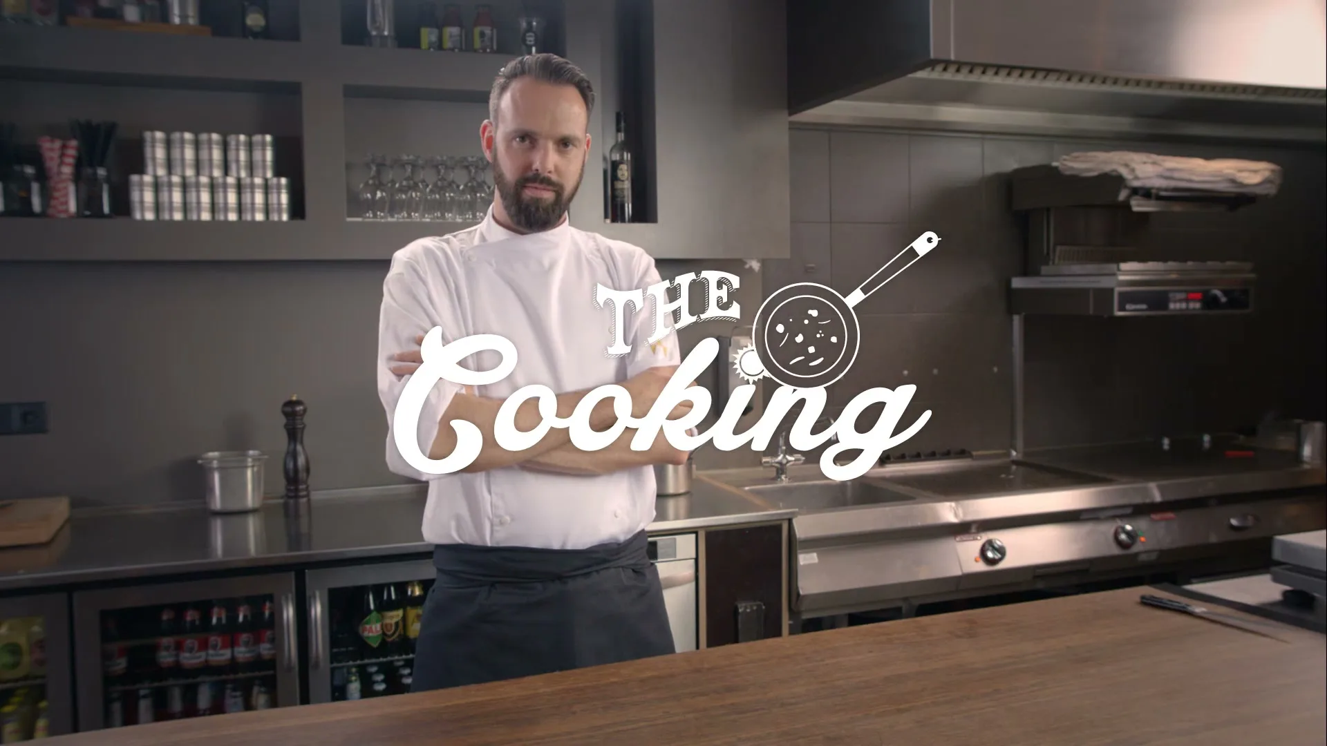 A male chef with a beard stands with his arms crossed in a professional kitchen, with "THE Cooking" text overlaid.