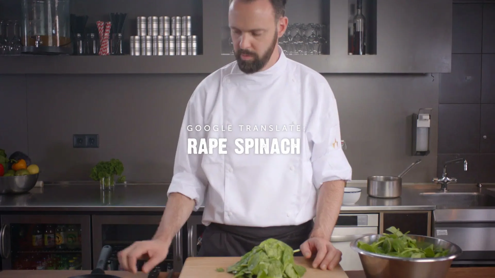 A male chef in a white uniform stands in a modern kitchen, looking down at a pile of spinach on a cutting board, then picks up a frying pan.