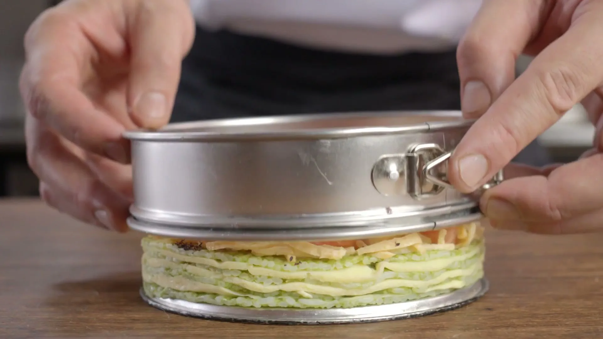 A chef's hands carefully remove a springform pan from a savory cake made of layered crepes, rice, salmon, avocado, and cherry tomatoes on a wooden table.