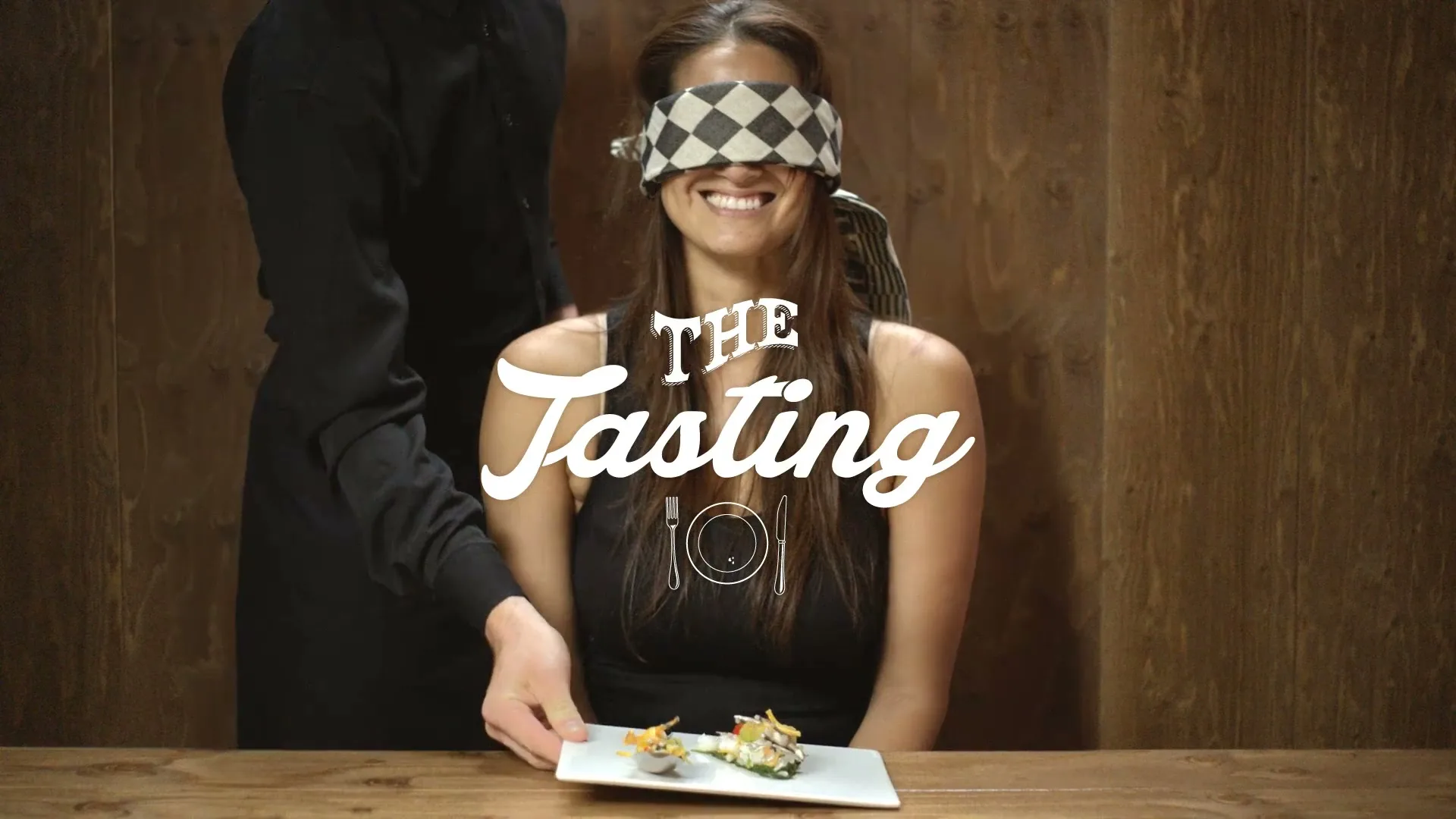 A blindfolded woman with a checkered blindfold smiles as a server in a black uniform places a plate of food in front of her on a wooden table.