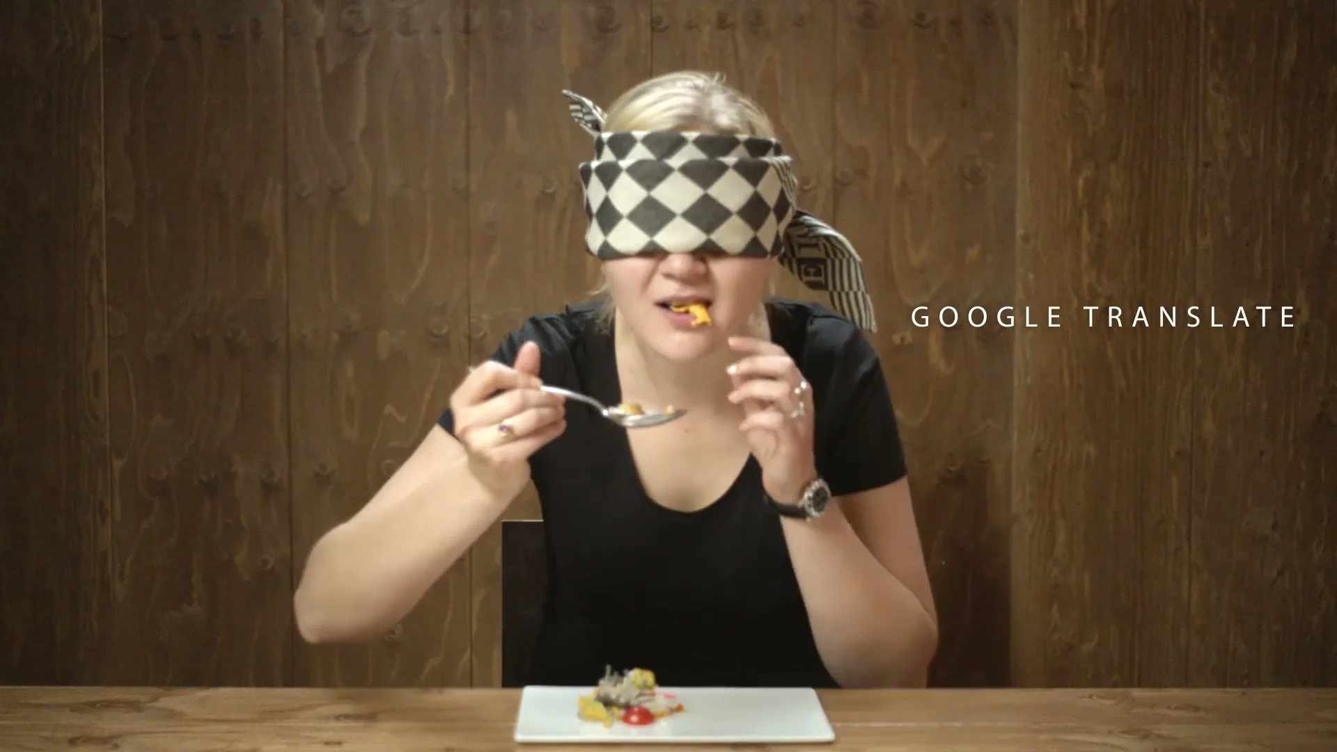 A blindfolded woman in a black t-shirt tastes a small portion of food from a spoon at a wooden table, with "GOOGLE TRANSLATE" text on the right.