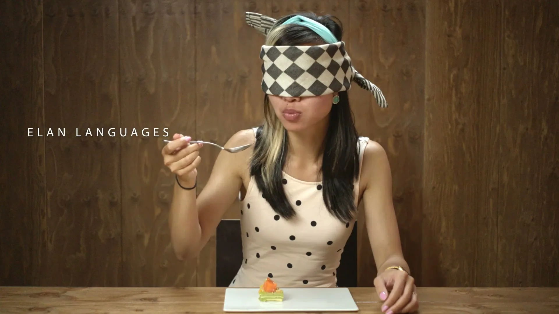 A young woman, blindfolded with a checkered cloth, sits at a wooden table, holding a spoon and tasting a small dish.