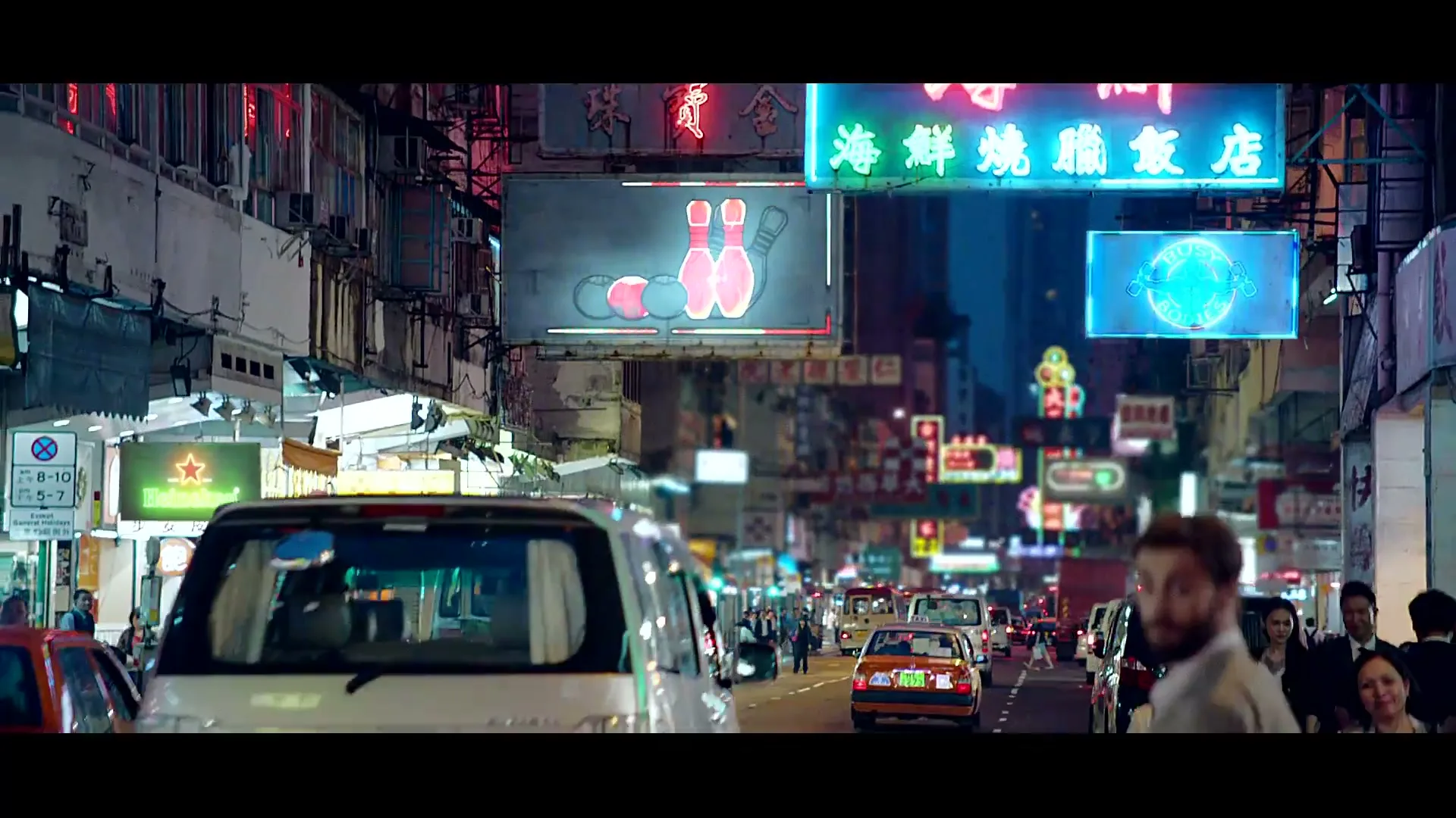A man in a beige jacket and blue pants walks through a busy city street at night, raising his hand to hail a taxi.