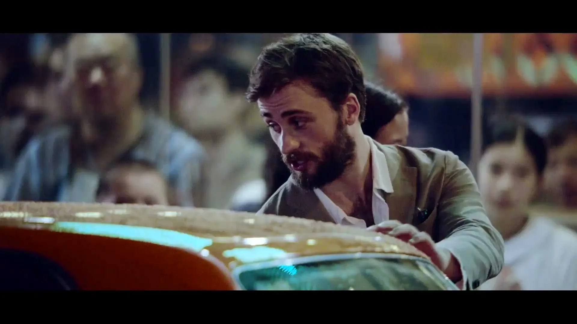 A man with a beard in a light jacket looks down at the back of an orange car, surrounded by a blurry crowd at night.