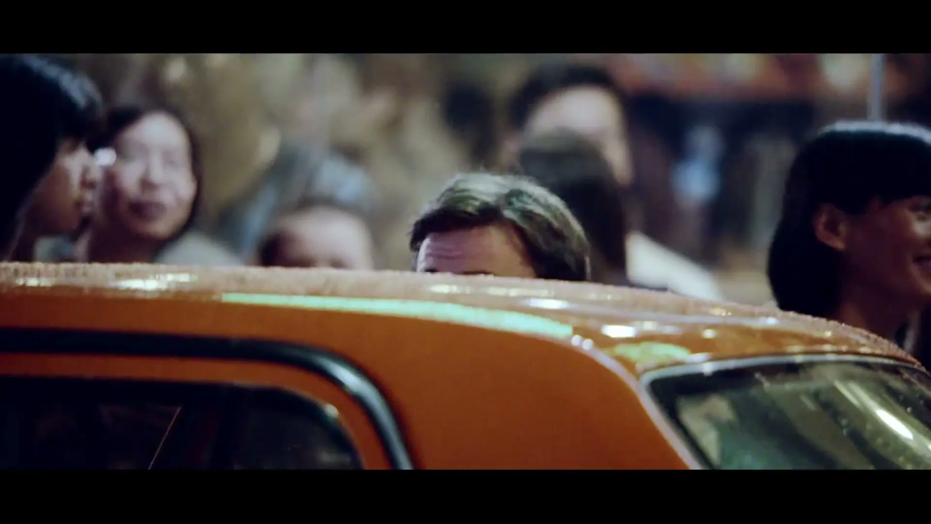 A man with a beard and a light brown jacket looks back at the camera from inside an orange taxi in a crowded city street at night.