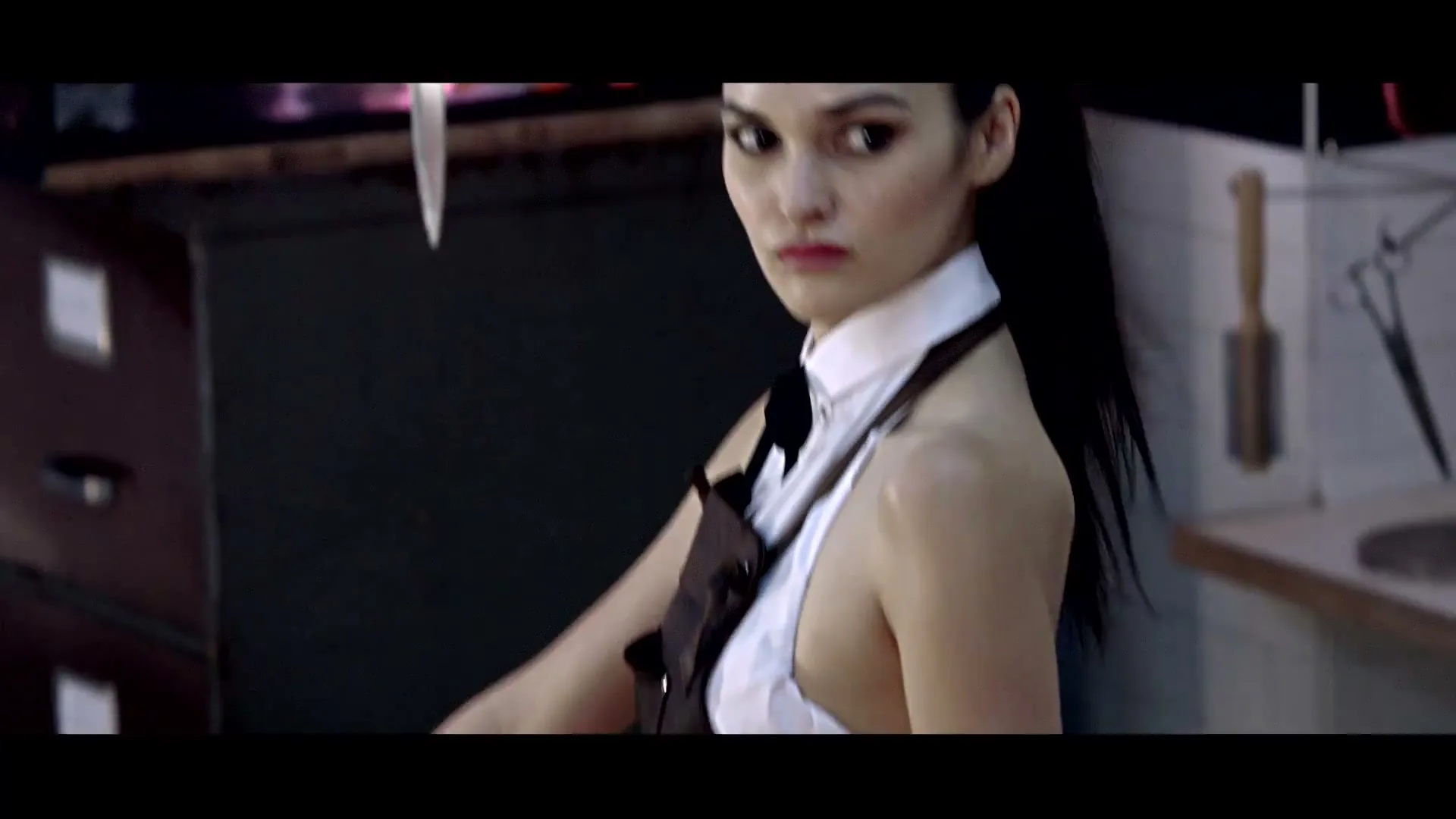A young woman with dark hair in a white shirt and brown apron looks over her shoulder with a serious expression in a dimly lit room.