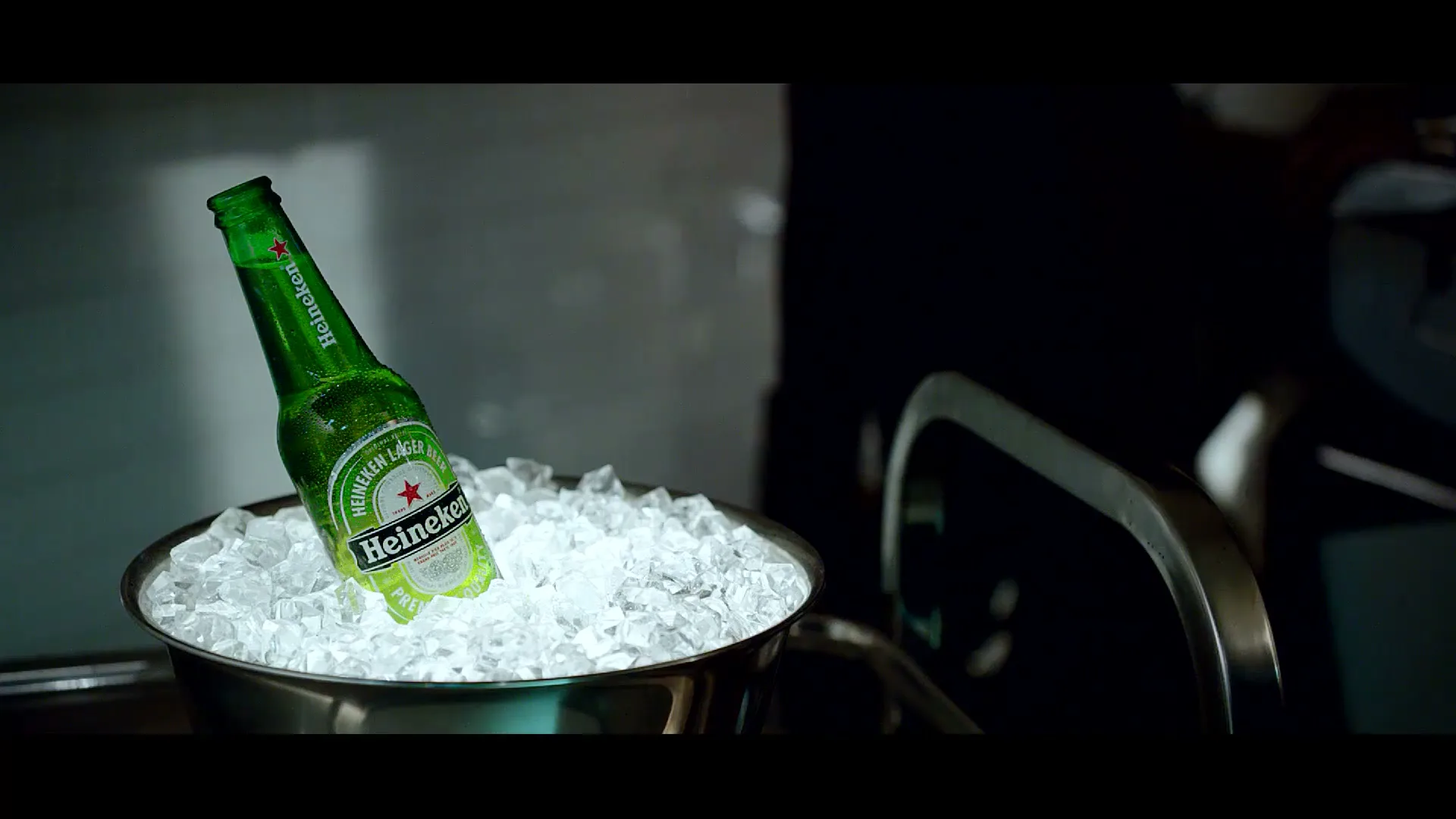 A green Heineken beer bottle sits in a silver ice bucket, with a dark background and a metallic handle visible on the right.