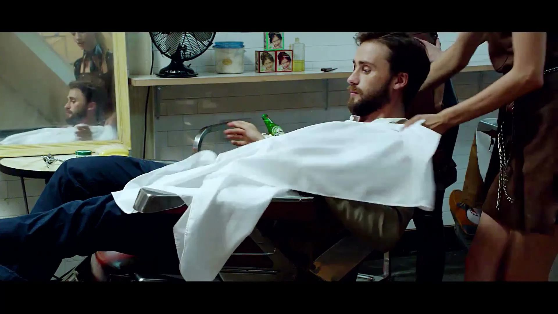 A man with a beard sits in a barber's chair as a woman drapes a white cloth over him in a dimly lit barbershop.