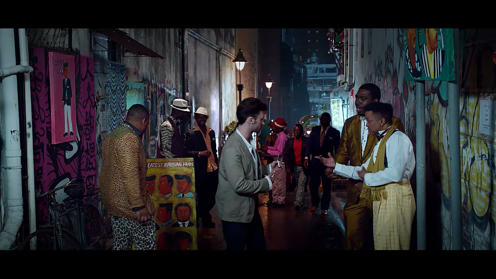 A young man in a light jacket stands in a graffiti-covered alley at night, interacting with several men in colorful, patterned suits.