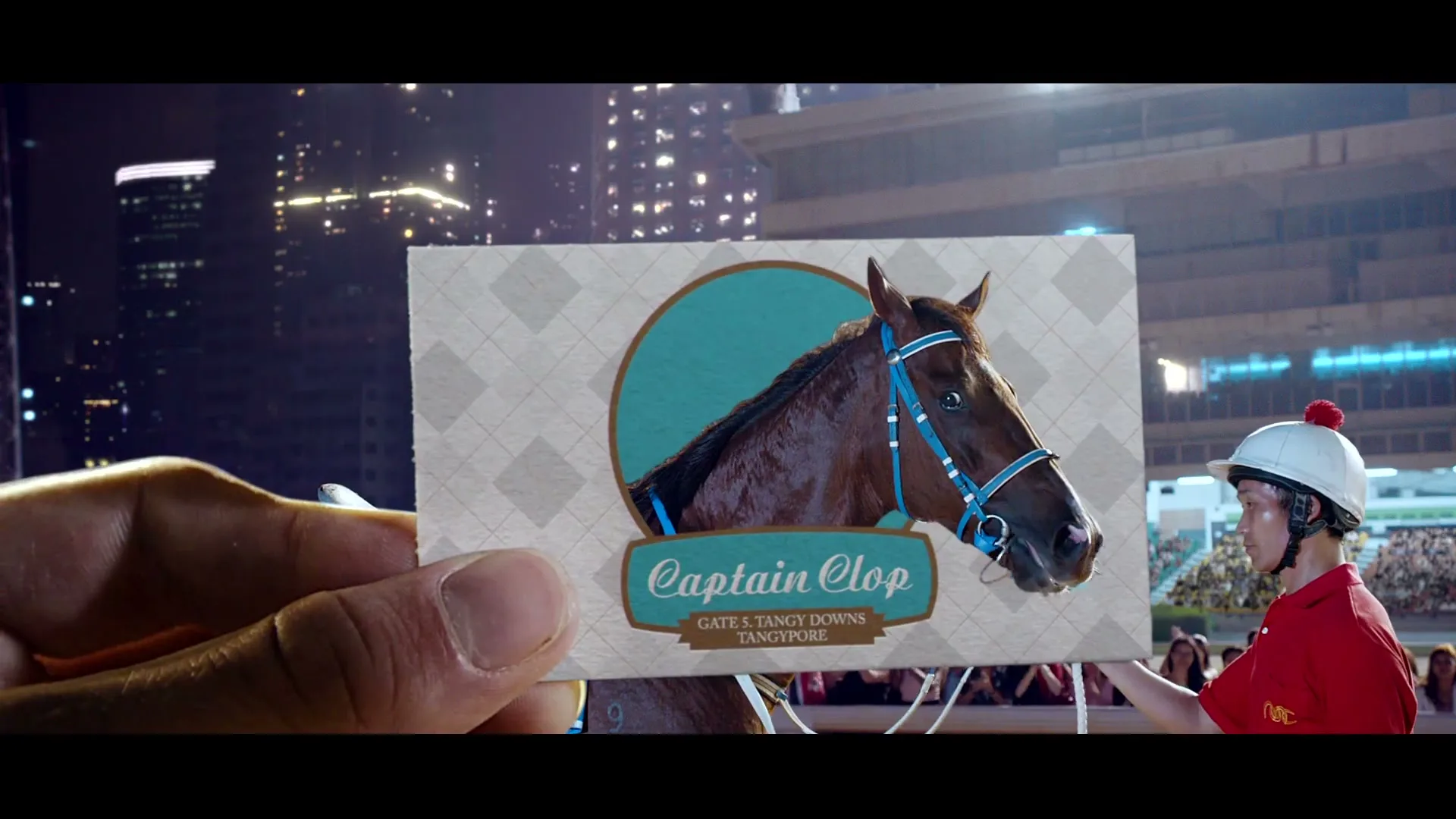 A man in a jockey helmet holds a card with a horse's picture, "Captain Clop" written on it, at a night horse race.