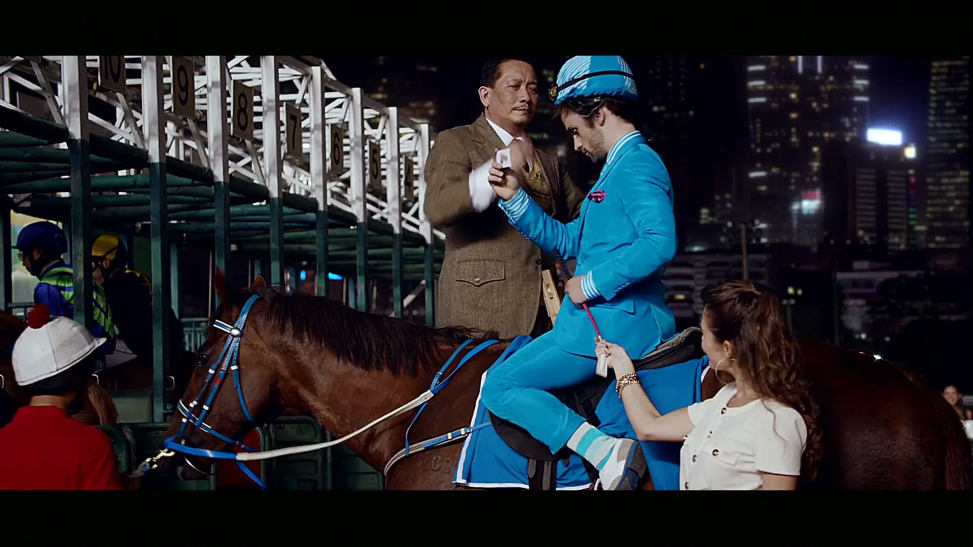 A man in a bright blue suit sits on a horse at a racetrack at night, while another man takes a photo and a woman adjusts his outfit.