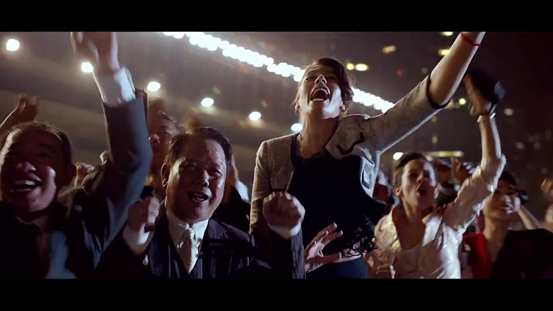 A diverse crowd of people, including a man in a suit and a woman in a jacket, cheer excitedly with their arms raised in an arena at night.