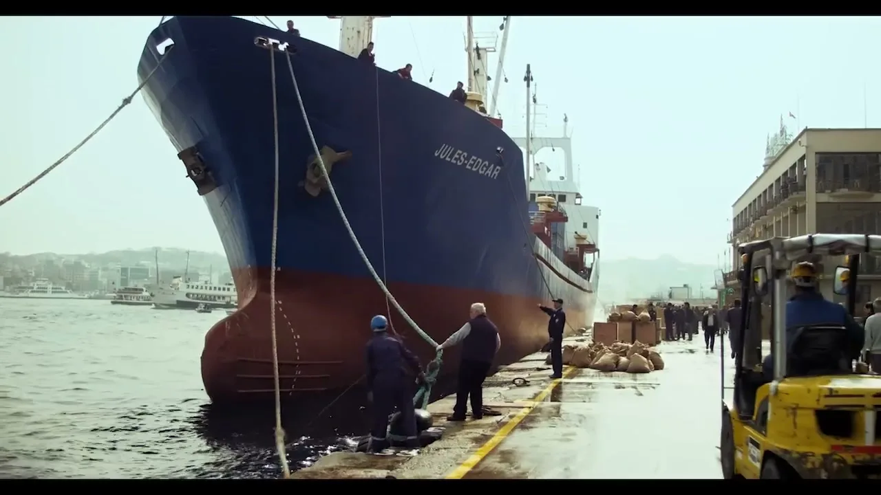 A large blue and red cargo ship named "JULES-EDGAR" is docked at a busy port, with several workers on the pier and a forklift moving cargo.