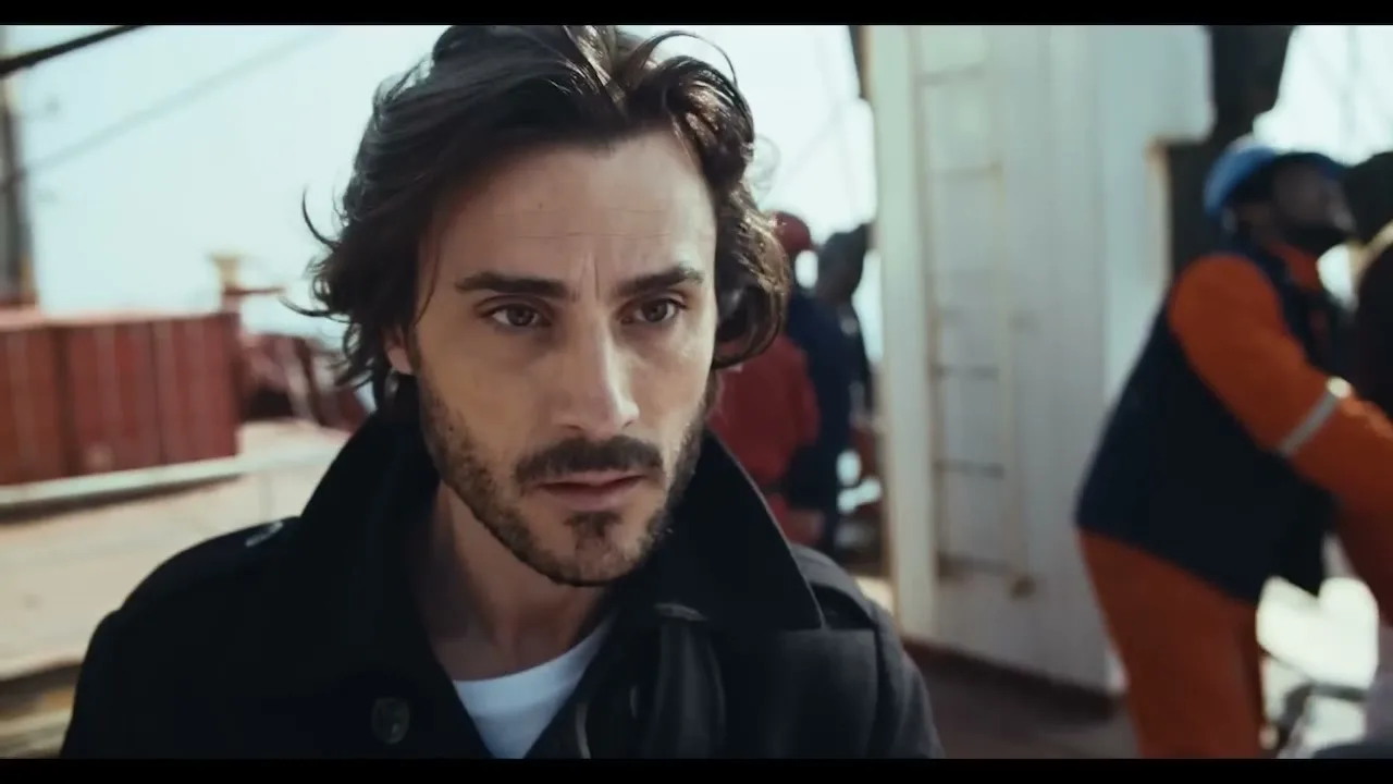 A man with dark hair and a beard looks up with a concerned expression on a ship's deck during the day.