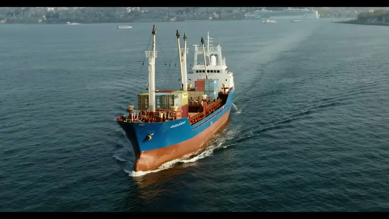 An aerial shot shows a cargo ship with containers sailing on a wide body of water, with a city skyline in the background under a clear sky.