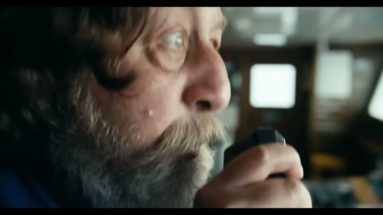 A bearded man with long hair, wearing a blue shirt, speaks into a walkie-talkie on a boat's bridge.