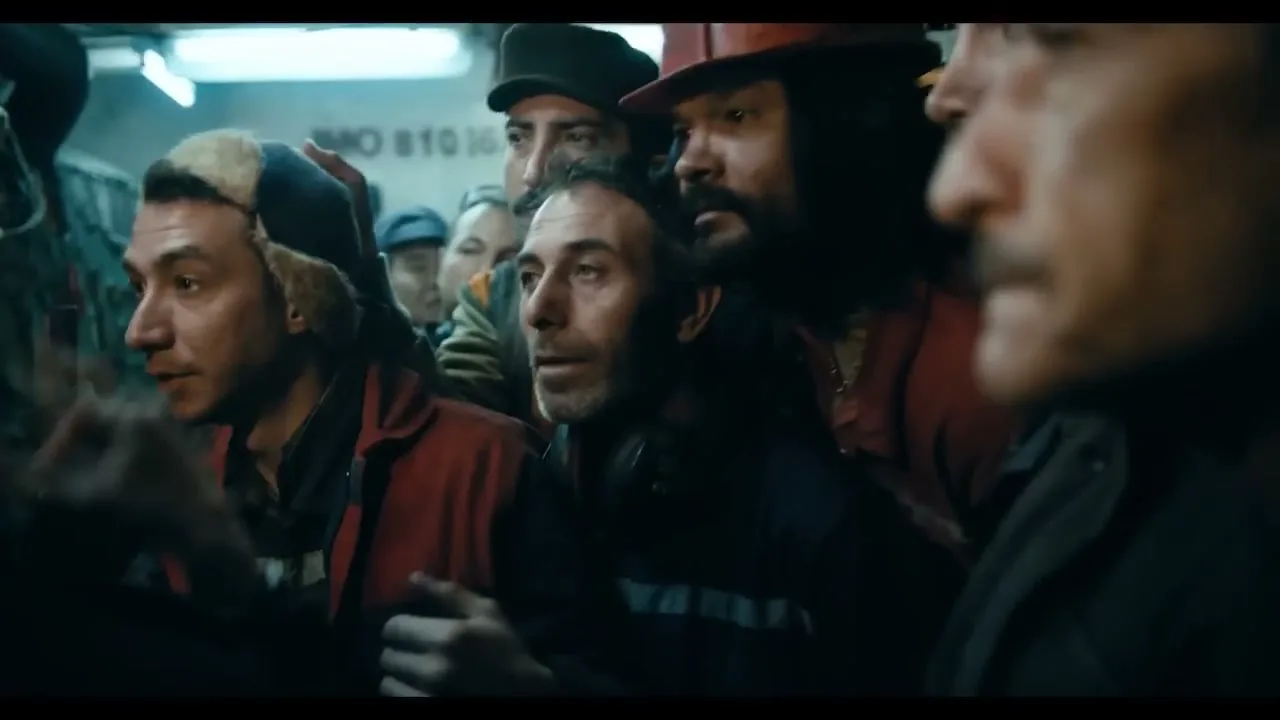 A group of men, some wearing hats, are tightly packed together in what appears to be a ship's interior, looking intently off-screen.