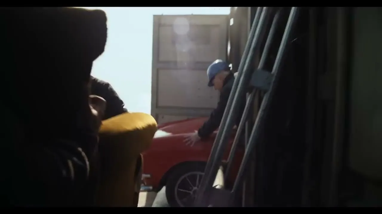 A man in a blue hard hat and dark jacket pushes a red vintage car out of a shipping container, seen from inside the container.
