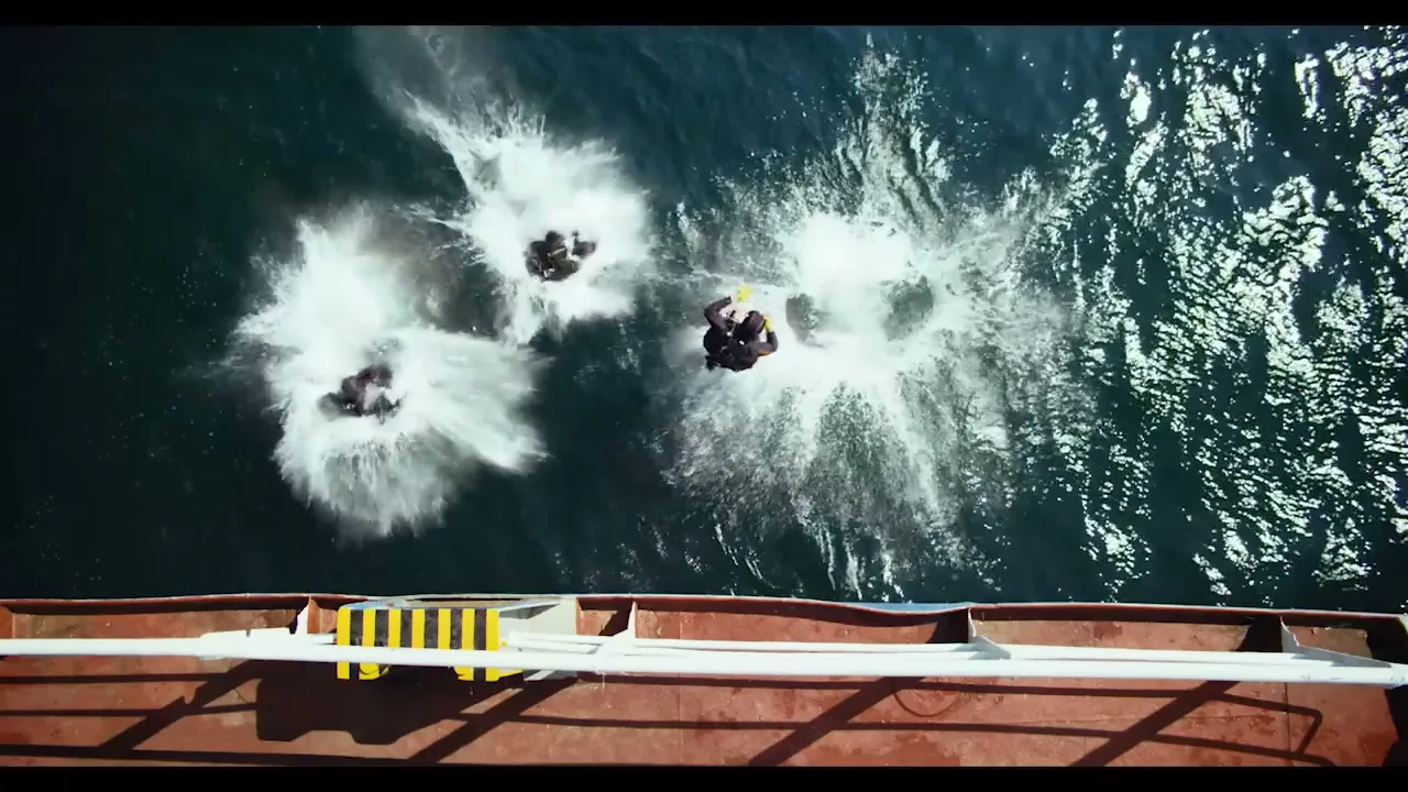 Three divers in black wetsuits jump from a ship into the ocean, creating large splashes in the bright sunlight.