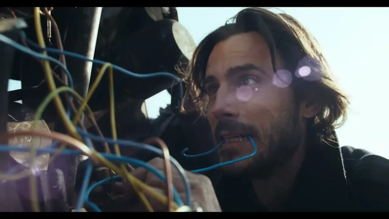 A man with long hair and a beard holds a blue wire in his mouth while working on a tangle of wires outdoors during the day.