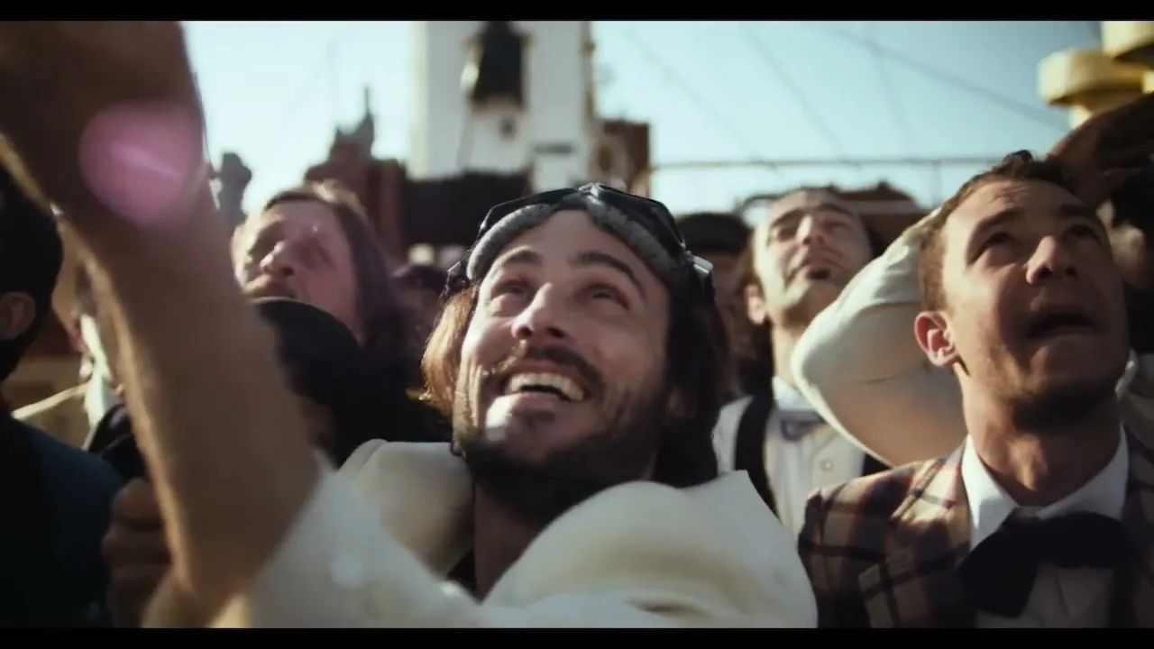 A crowd of people, including a man with a beard and goggles, look up with joyful expressions on a sunny day.