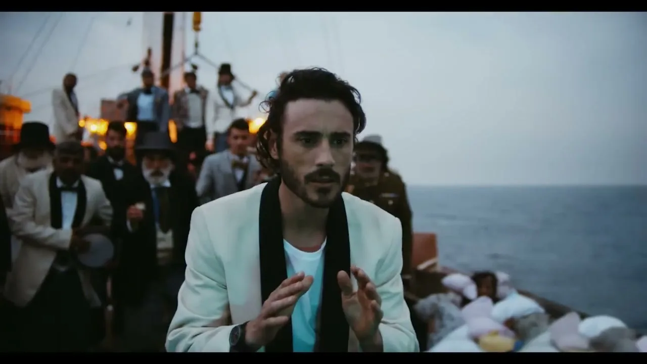 A man in a white tuxedo jacket looks concerned while standing on a boat with a crowd of men in formal wear behind him, at sea.