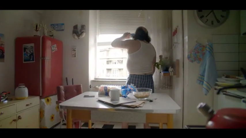 A man in a white tank top and plaid boxers stands at a kitchen window, looking out and drinking from a bottle, in a messy kitchen.