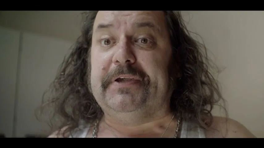 A middle-aged man with long, curly hair and a mustache looks directly at the camera with a surprised expression, wearing a white tank top indoors.