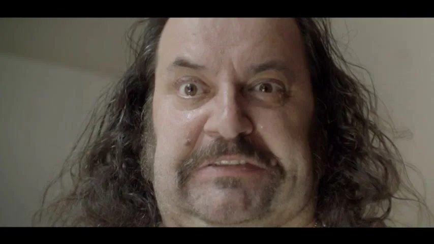 A man with long dark hair and a mustache looks directly at the camera with wide, angry eyes, his face slightly sweaty.