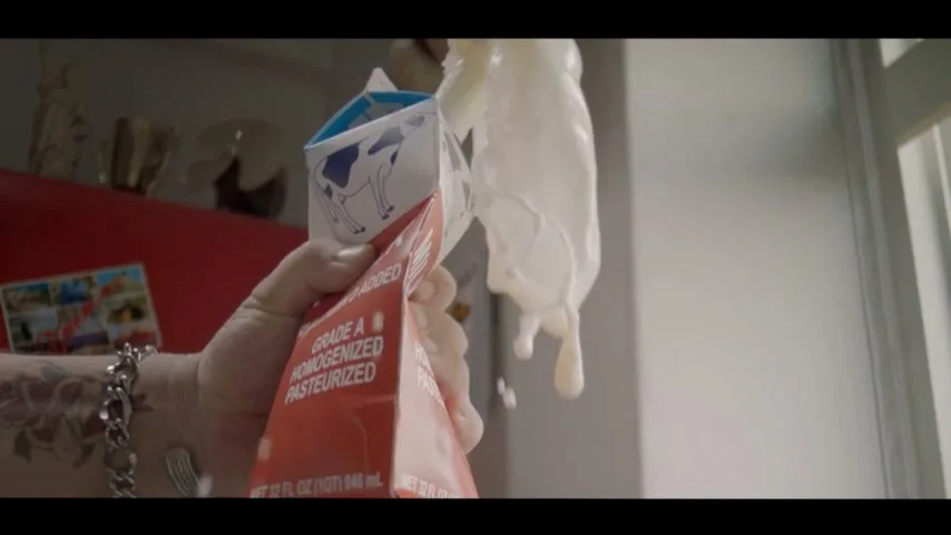 A person's tattooed hand squeezes a carton of milk, causing milk to spray out and splash against a white wall in a kitchen.