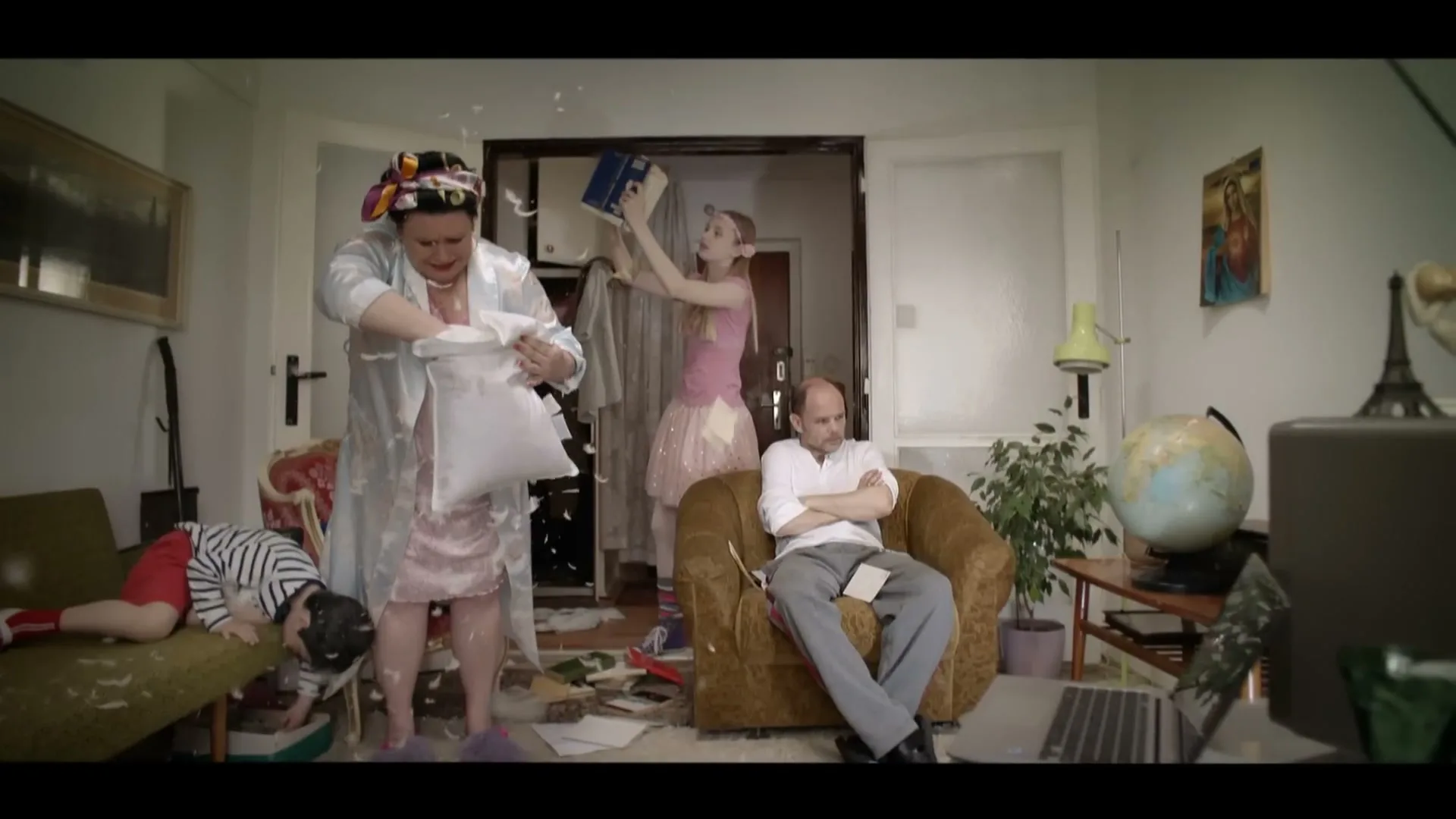 A family of four is in a messy living room, with a woman tearing a pillow, a girl throwing clothes, a boy lying on a couch, and a man sitting in an armchair.