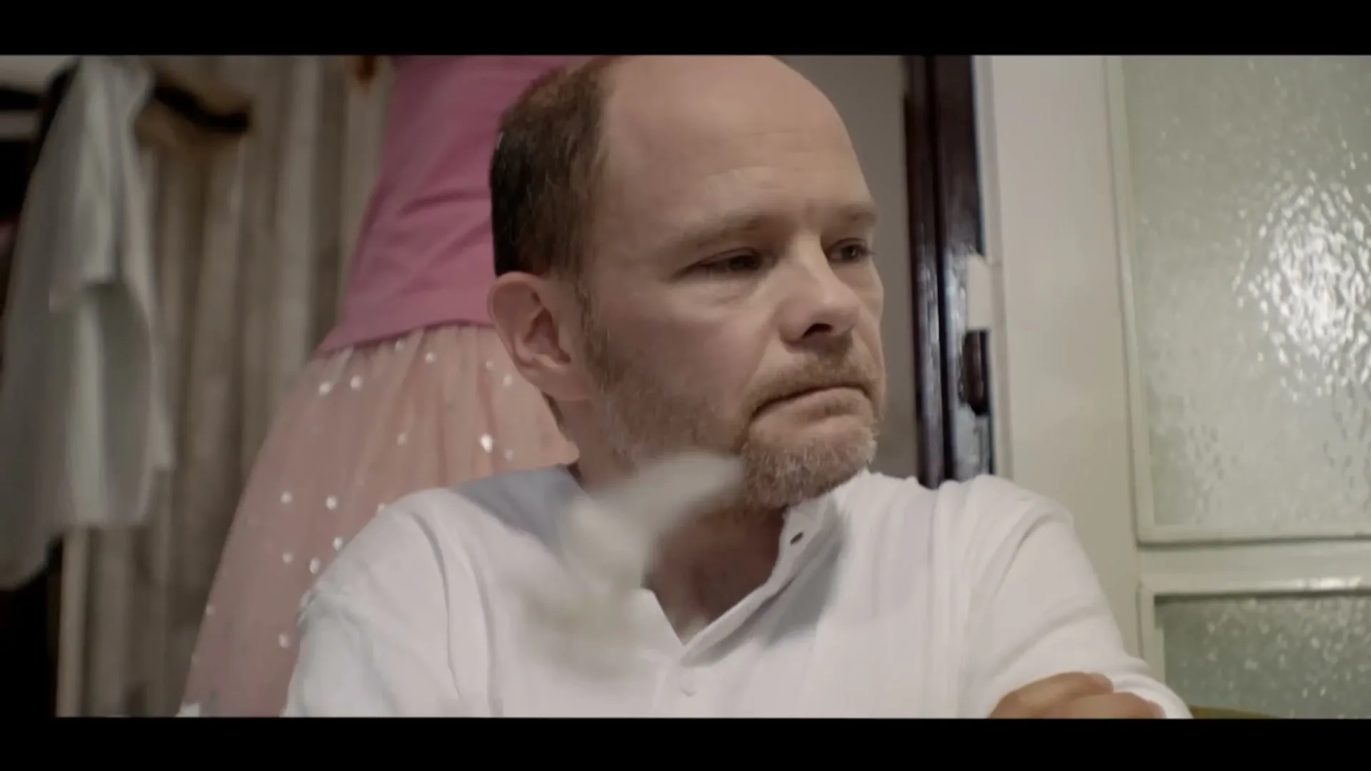 A middle-aged man with a beard sits looking sad, while a woman in a pink top and sparkly skirt stands behind him, in a domestic interior.