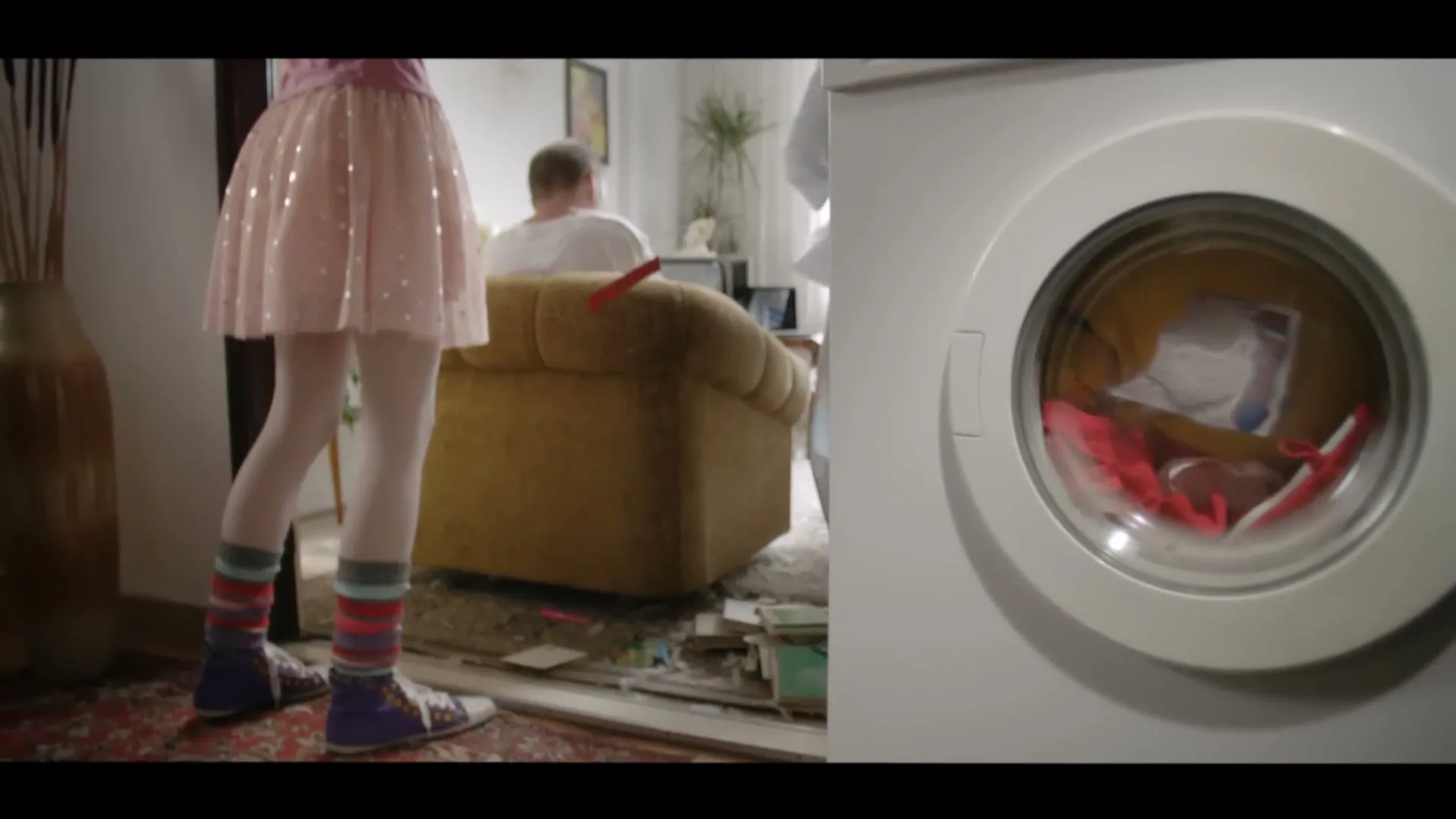 A split screen shows a young girl in a pink skirt and colorful socks standing in a messy living room, while a washing machine tumbles clothes on the right.