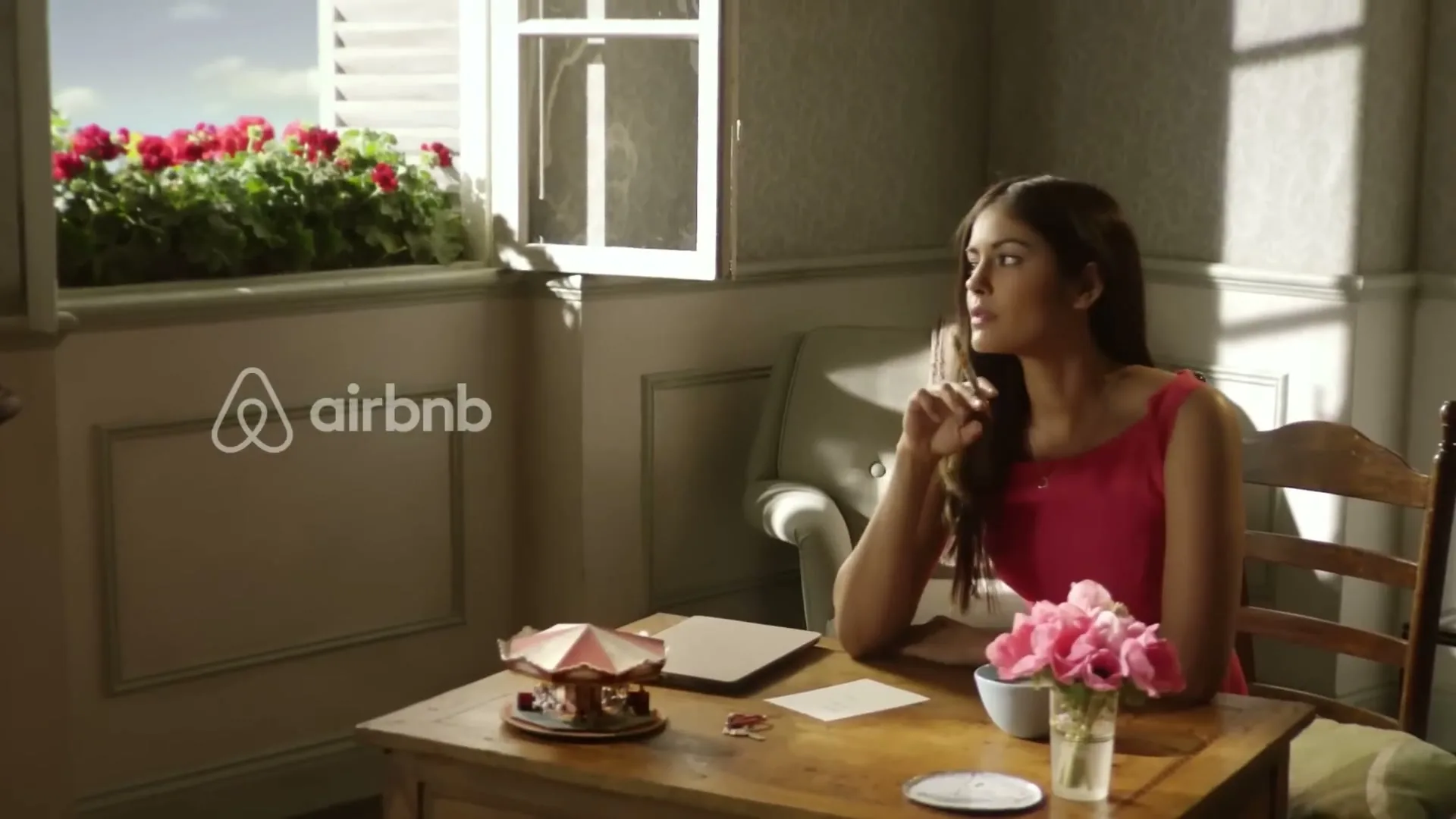 A young woman in a pink dress sits at a wooden desk, looking out an open window with red flowers, in a sunlit room.