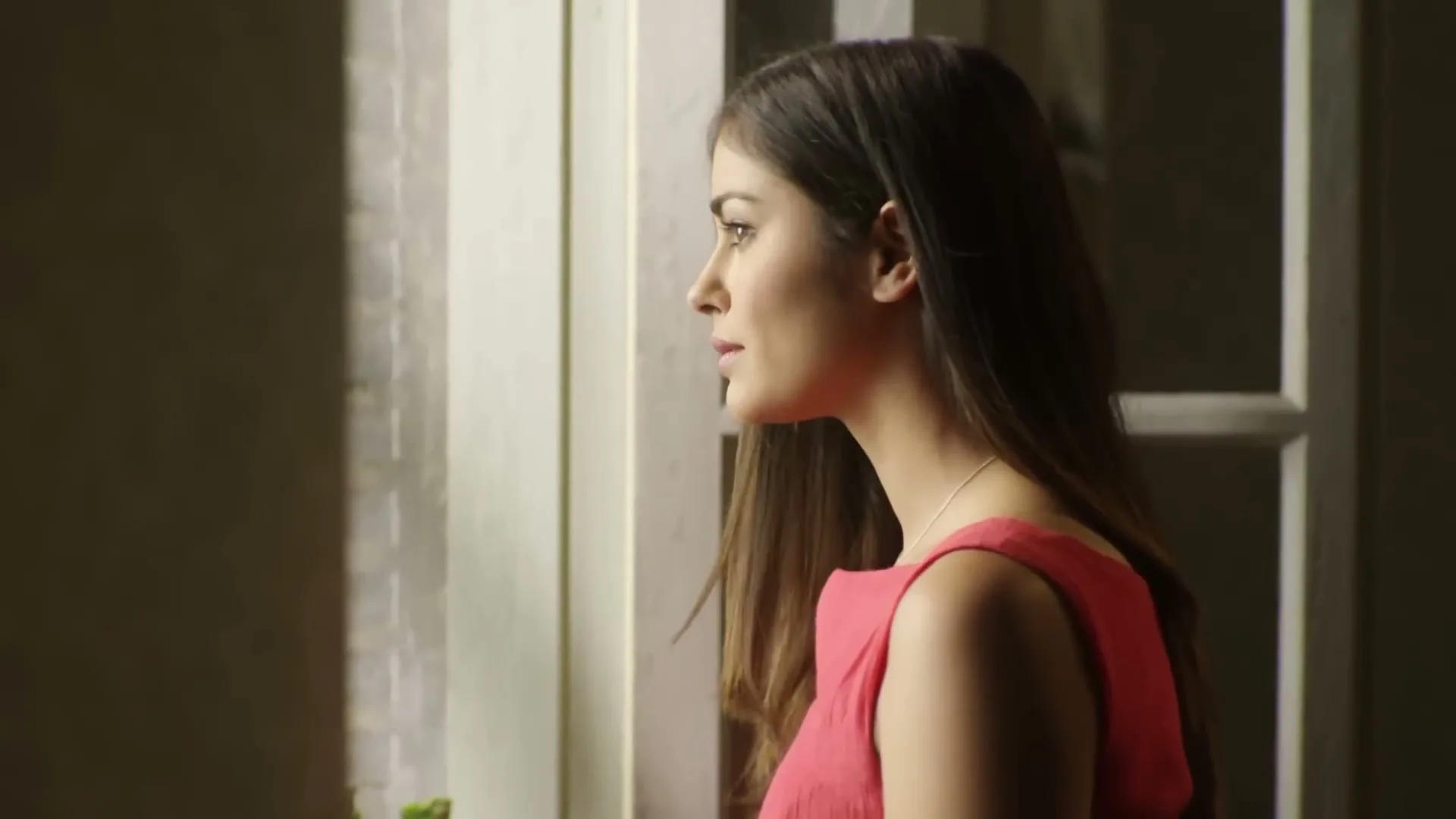 A young woman with long brown hair, wearing a pink dress, looks out a window in a softly lit room.