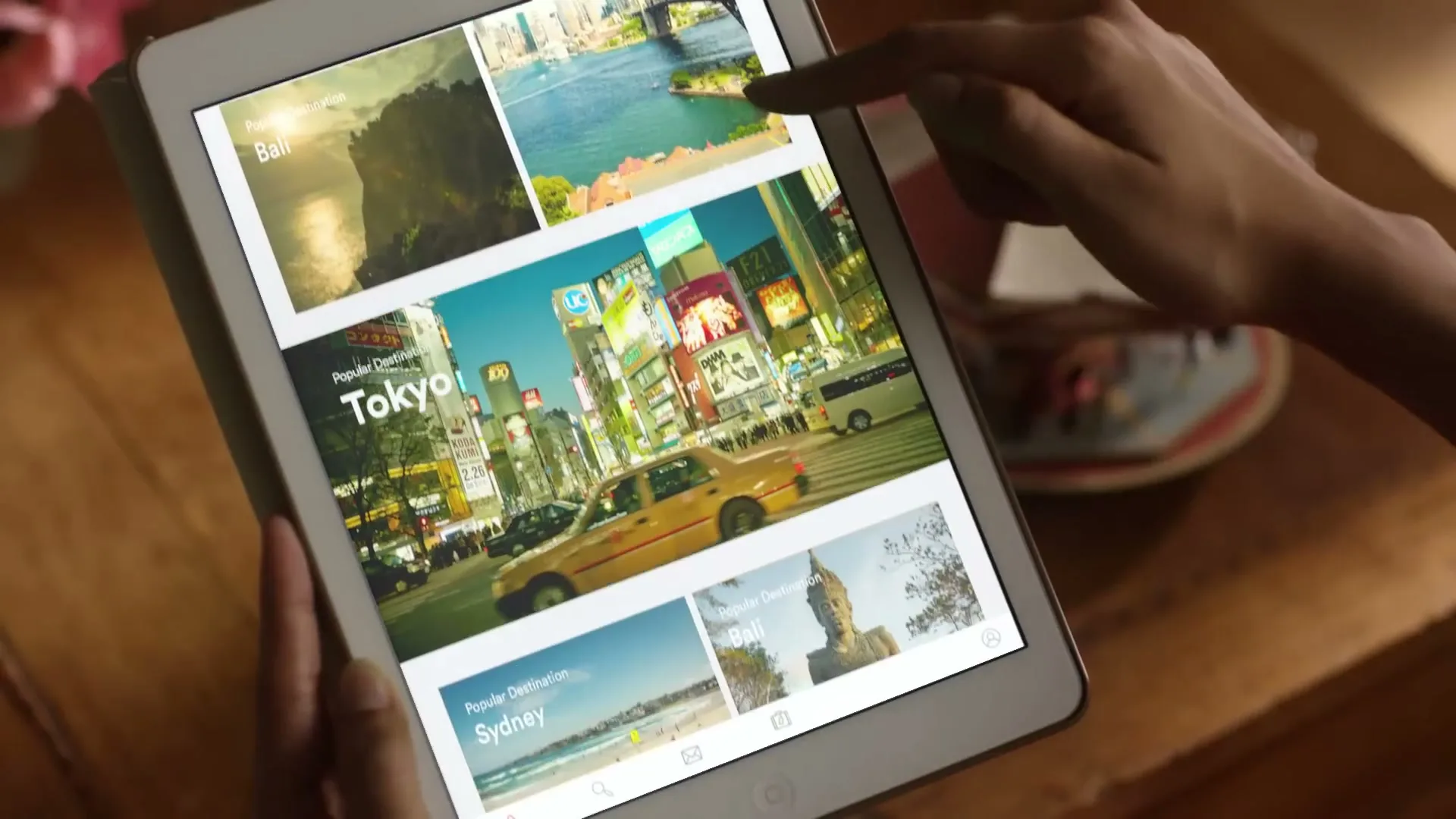 A person's hands hold a tablet displaying travel destinations, with a finger scrolling through options on a wooden table.