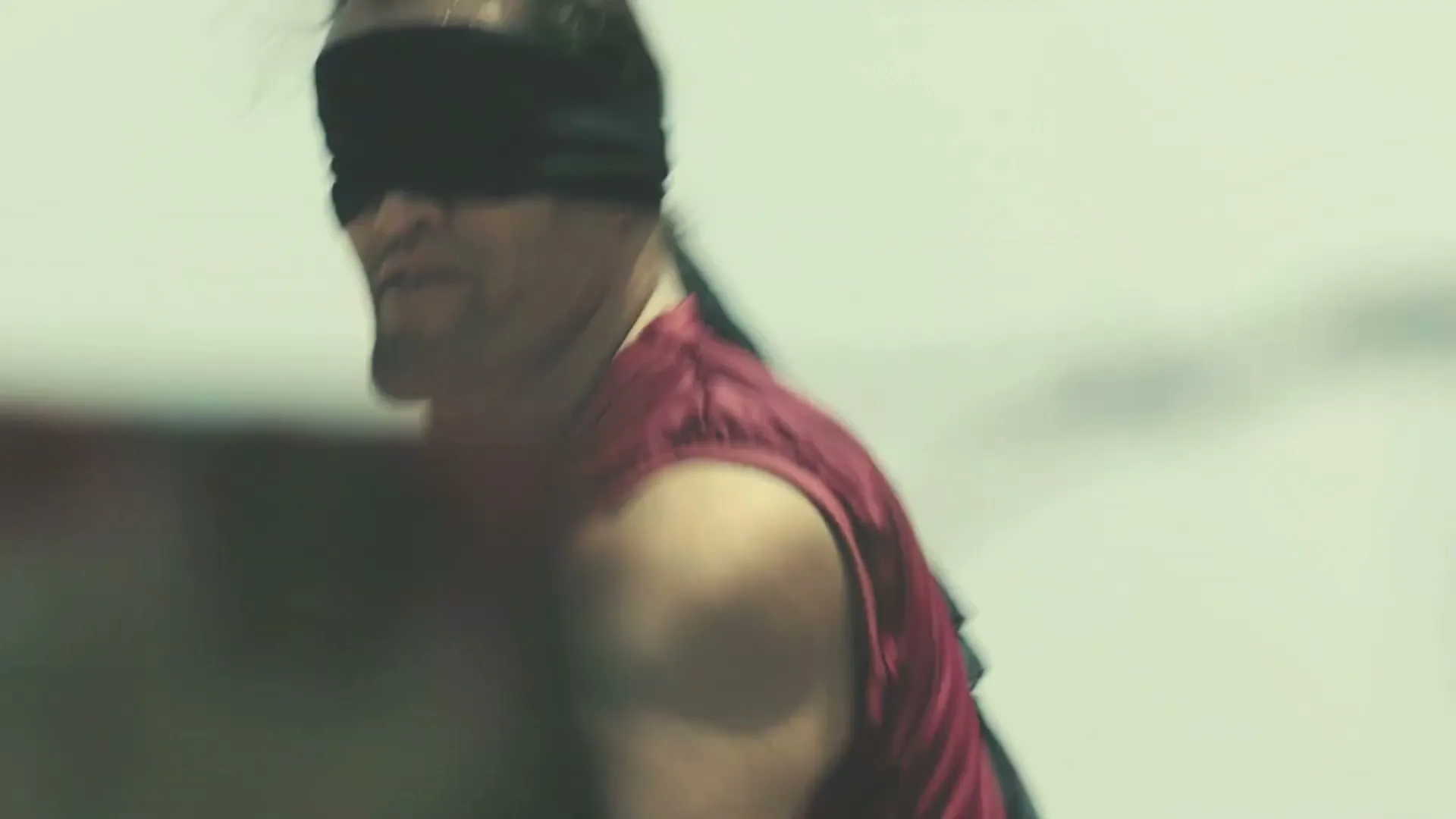 A blindfolded man in a red tank top runs, looking disoriented, as children play in the background on a sunny day.